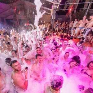 A group of partygoers covered in foam, laughing and dancing under neon lights at the Flirt Foam Party in Aria Complex, Malta.