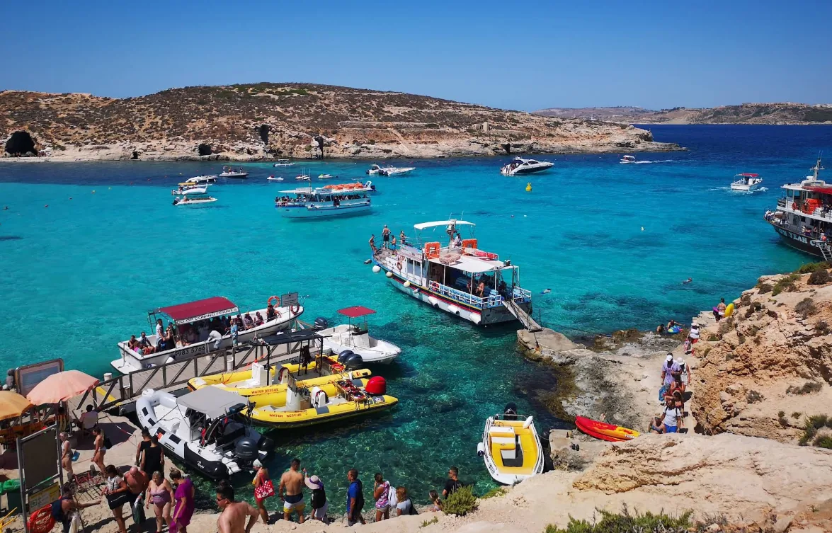 Traghetti colorati, motoscafi e gommoni ormeggiati nella Blue Lagoon Comino Malta con folla estiva sulla costa rocciosa calcarea sotto cielo blu intenso