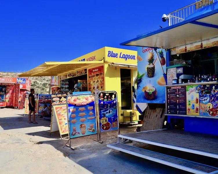 Chiosco di cibo Blue Lagoon colorato giallo e blu su Comino Malta con menu, segnaletica cocktail ananas e stand gastronomici sotto un cielo blu profondo