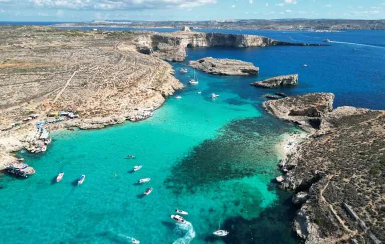 Aerial drone panorama of Blue Lagoon Comino Malta showing scattered sailboats and motorboats in electric turquoise water with white limestone cliffs and Comino Tower in the background