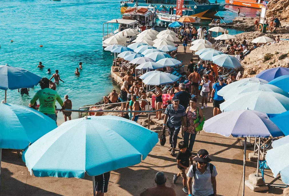 Folla densa di turisti in alta stagione che camminano e siedono sotto parasoli blu e bianchi lungo la passeggiata della Blue Lagoon Comino Malta con acqua turchese e traghetti ormeggiati sullo sfondo