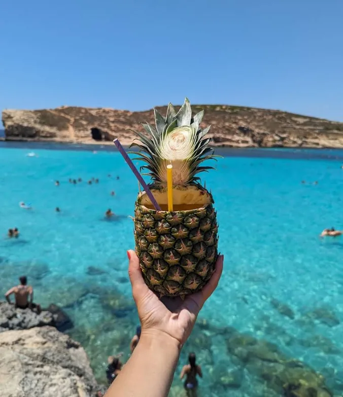 Mano che regge un cocktail all'ananas fresco in primo piano con l'acqua turchese cristallina della Blue Lagoon Comino Malta e nuotatori visibili sullo sfondo sotto un cielo blu profondo
