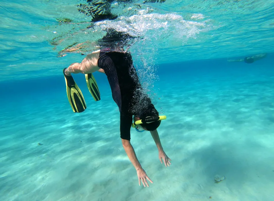 Foto subacquea di uno snorkellista in muta nera con pinne gialle che si tuffa a testa in giù nell'acqua turchese cristallina sopra il fondale sabbioso della Blue Lagoon Comino Malta