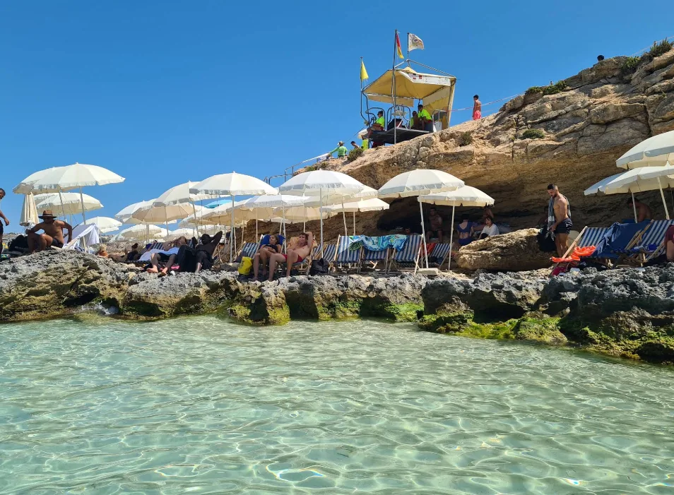 Vista dall'acqua con file di lettini bianchi e parasoli sulle rocce piatte della Blue Lagoon Comino Malta con torre di salvataggio e acqua cristallina poco profonda in primo piano