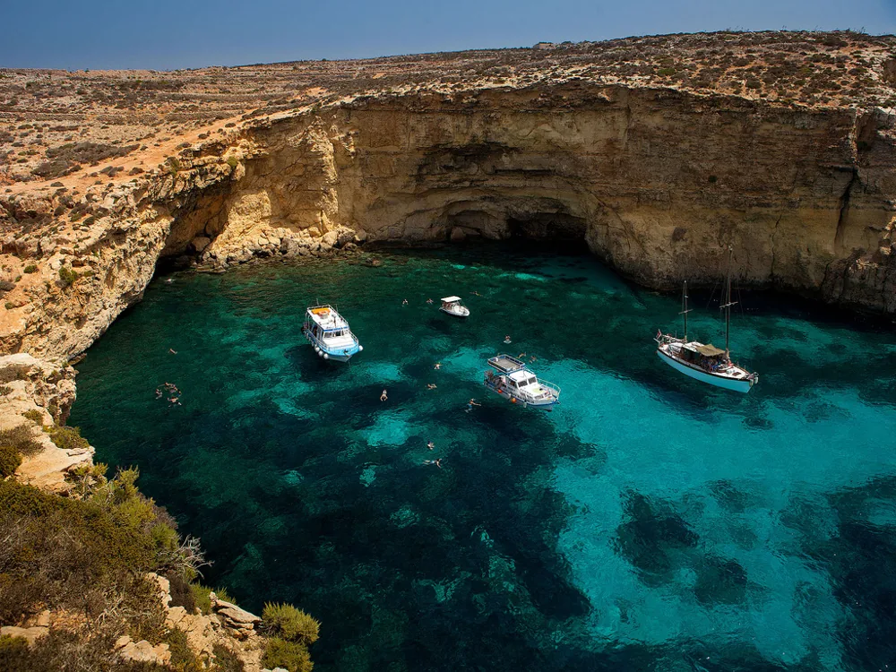 Vista aerea dalla scogliera del Crystal Lagoon Comino Malta con due yacht a motore bianchi e una barca a vela ancorati in acque smeraldo e turchesi profonde incorniciate da scogliere calcaree dorate e un arco roccioso naturale