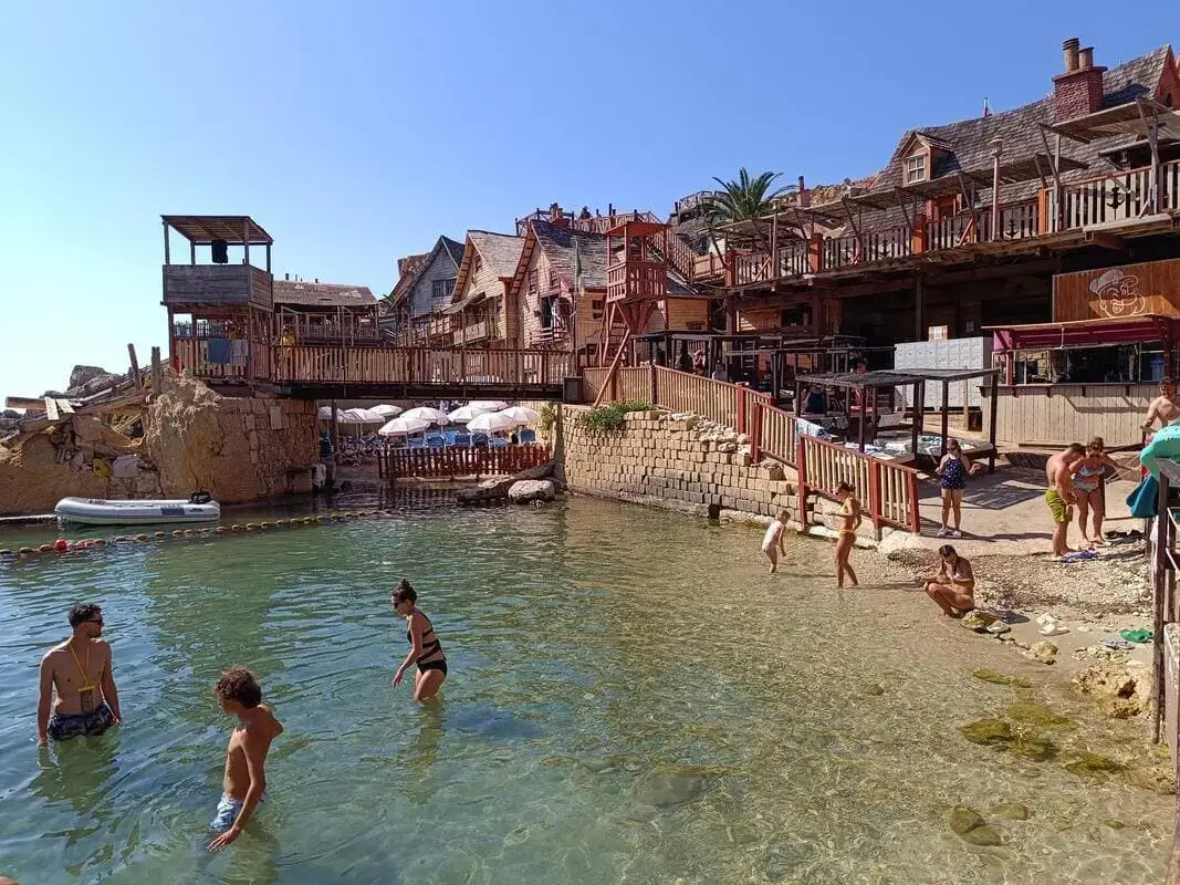 Spiaggia Popeye Malta - Zona nuoto Anchor Bay Visitatori che nuotano nell\'acqua limpida di Anchor Bay al Popeye Village Malta con gli edifici in legno del Villaggio Popeye sullo sfondo